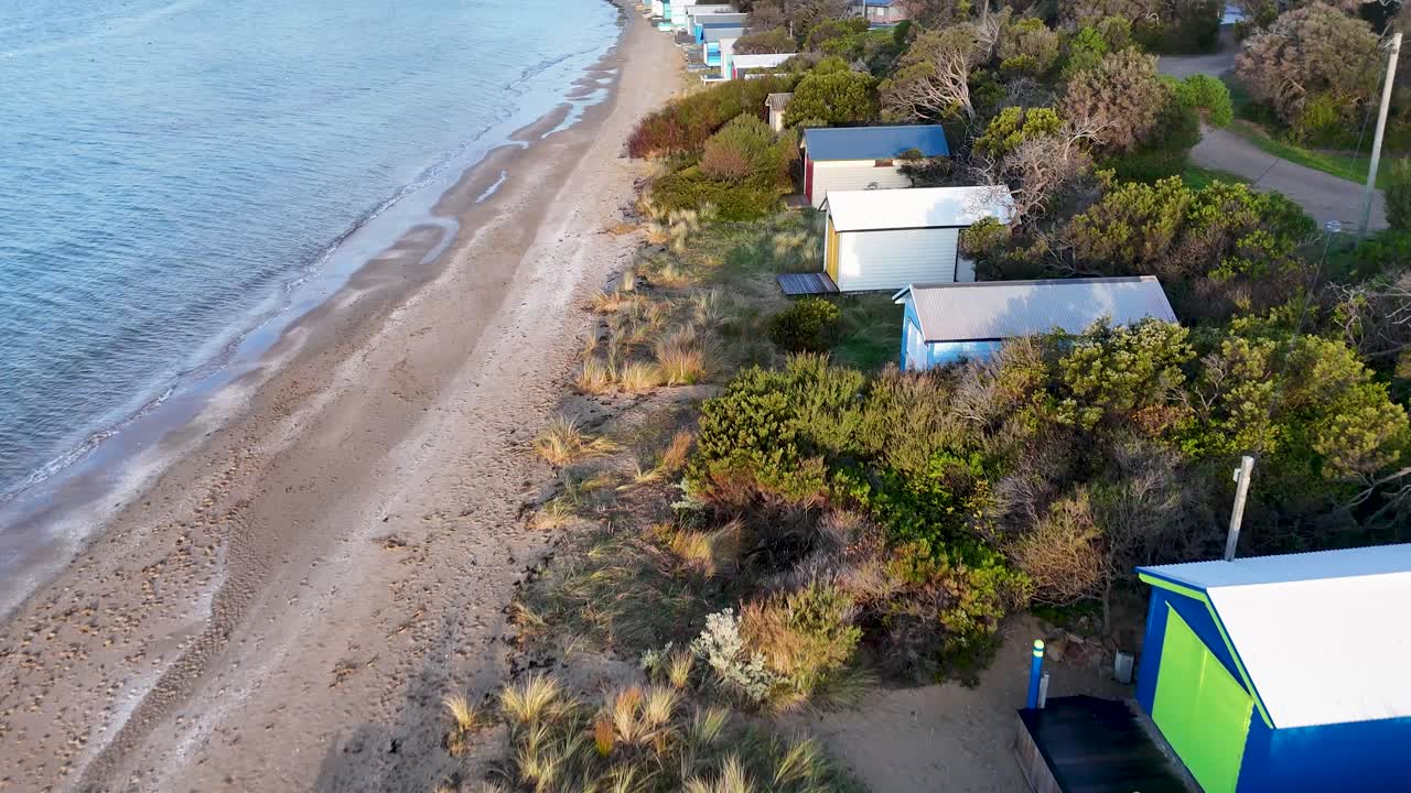 Drone captures person walking dog by colorful beach huts, soft daylight, gentle movement, coastal scenery