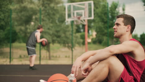 Im-Vordergrund-Ein-Müder,-Gutaussehender-Basketballspieler,-Der-Eine-Pause-Macht,-Während-Er-Auf-Dem-Boden-Sitzt-Und-Wasser-Auf-Einem-Basketballplatz-Im-Freien-Trinkt