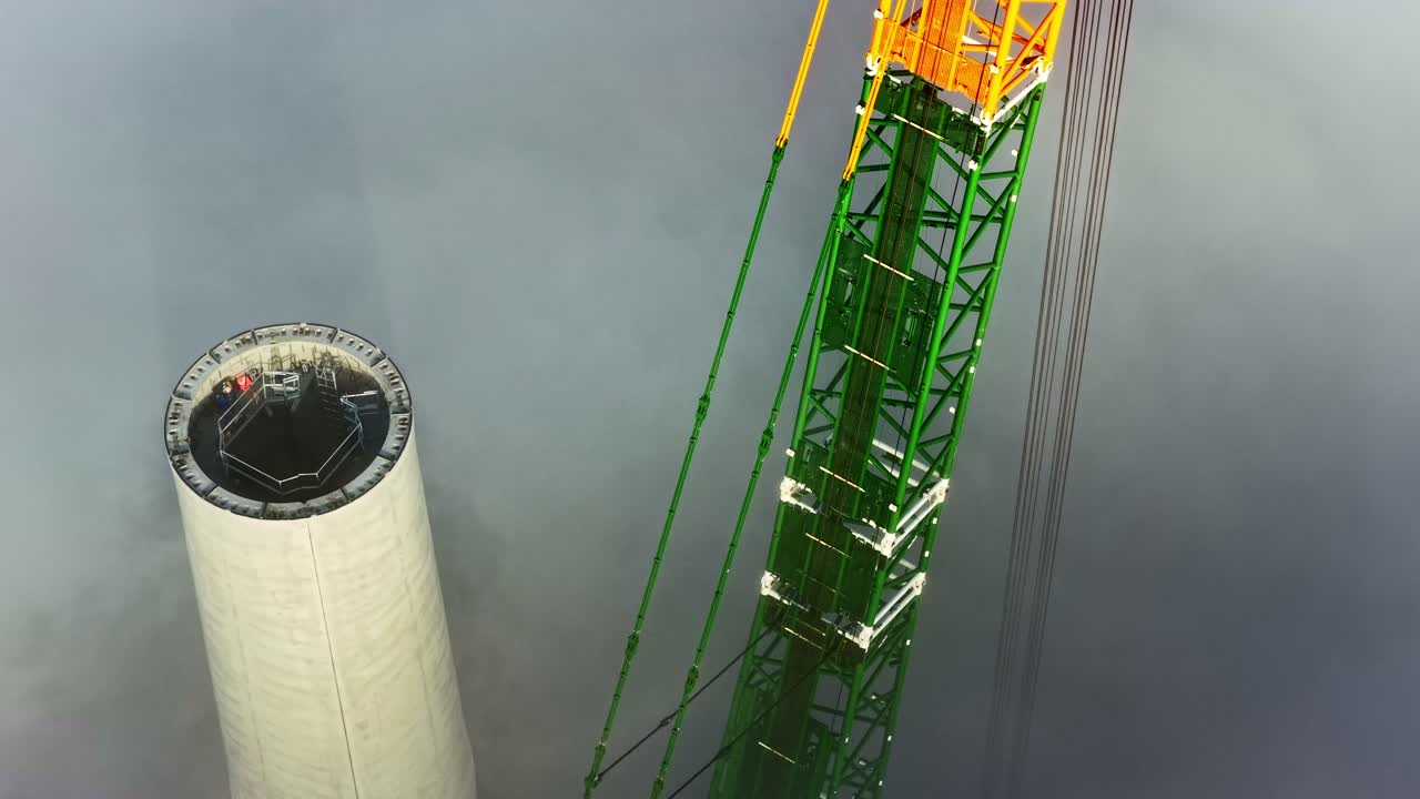 Dramatic aerial parallax reveals a massive construction crane assembling a huge wind turbine tower for renewable energy, shrouded in atmospheric clouds in Latvia
