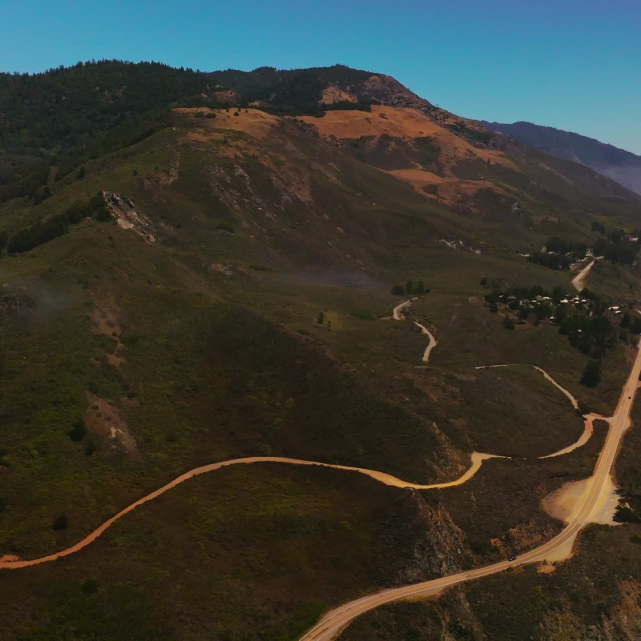 Roads passing through uneven mountainous landscape of California coastline. Pine tree forest overgrowing the mountain tops. Aerial perspective