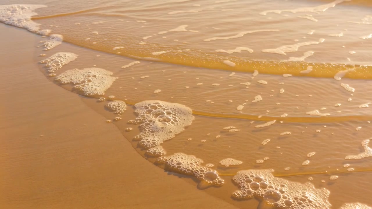 Fronte di onda spumeggiante che avanza sulla sabbia bagnata, mentre la marea spinge una pellicola d'acqua e ammassi di schiuma.