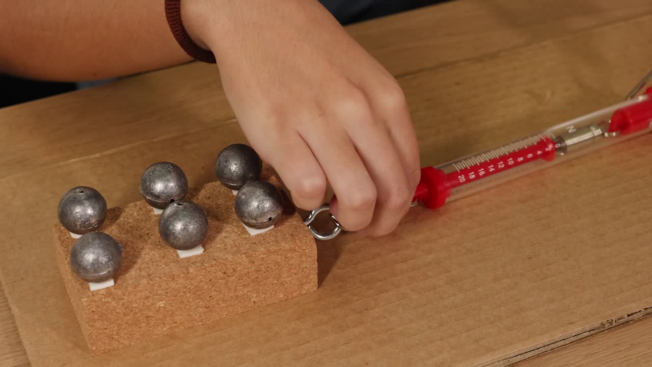 A hand pulls a weighted block on cardboard using a spring scale, illustrating friction and force dynamics