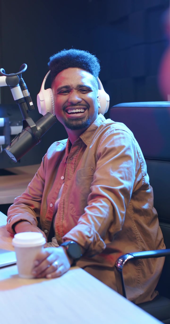 Video verticale: Uomo afroamericano che ride nel microfono in studio con le cuffie dopo una battuta del co-conduttore