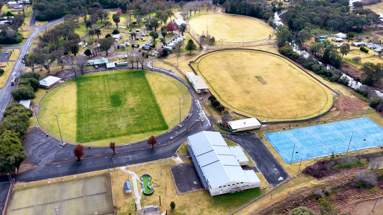 Drone footage glides above sports fields, tennis courts, and community buildings in a small Australian town, under bright, natural daylight with steady camera movement