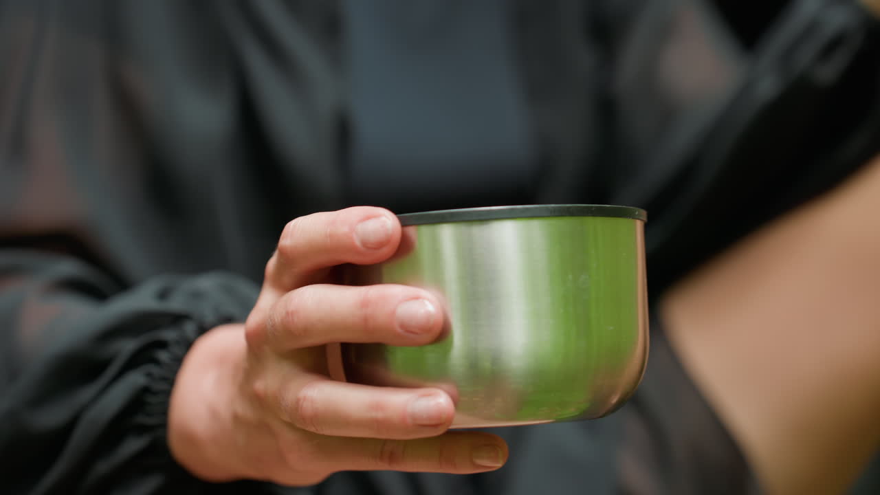 Close up of young woman dressed in black outfit sipping juice from stainless flask cover while holding pastry in other hand, soft natural light highlighting gesture, calm atmosphere