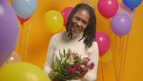 studio portrait of woman wearing birthday headband holding bunch of flowers celebrating with balloons