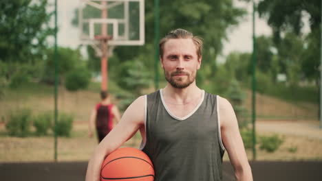 Retrato-De-Un-Apuesto-Jugador-De-Baloncesto-Rubio-Barbudo-Sosteniendo-Una-Pelota,-Mientras-Mira-La-Cámara-Y-Sonríe-Con-Confianza-En-Una-Cancha-De-Baloncesto-Al-Aire-Libre-1