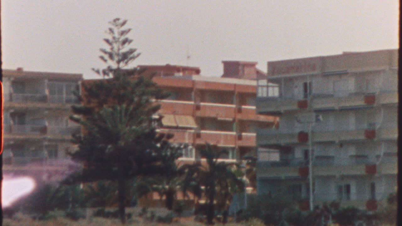Vintage vibes emanate from this zooming shot of a hotel complex, complete with a prominent pine tree, likely located in Spain during the 1970s