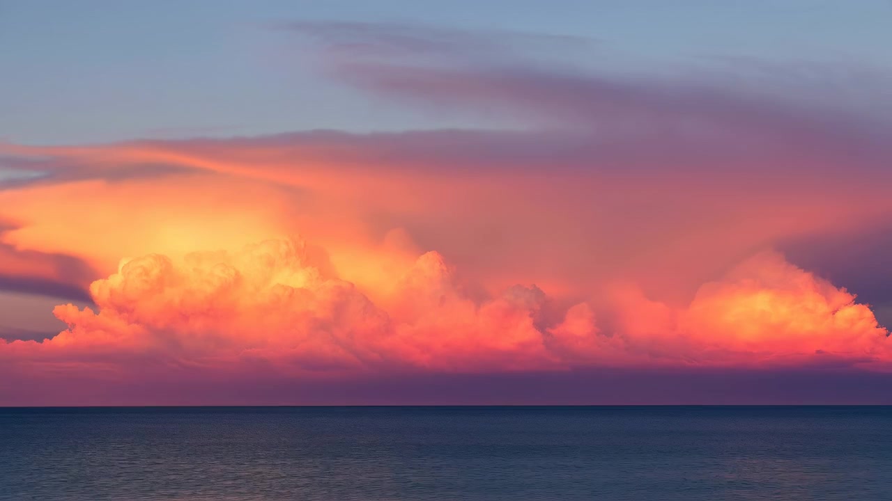 広大な海の景色は、劇的な低い雲の帯の下に穏やかな海が広がり、暖かい日の出の光を受けて雲の頂が珊瑚色からオレンジ、ピンクへと輝きを変え、穏やかな雰囲気を作り出しています。