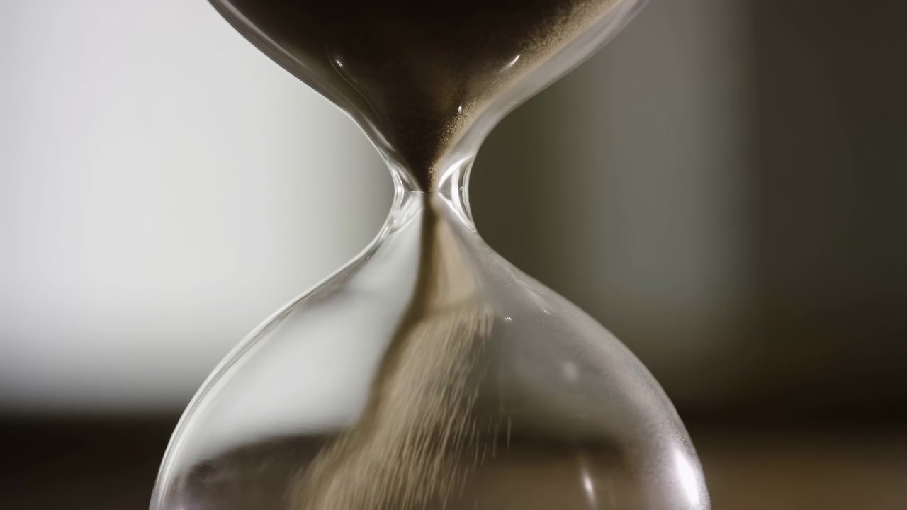 Clessidra di vetro in primo piano con sabbia fine marrone che scorre attraverso il collo stretto, formando un soffice mucchio sotto mentre la camera superiore si svuota lentamente in una luce da studio calma e neutra