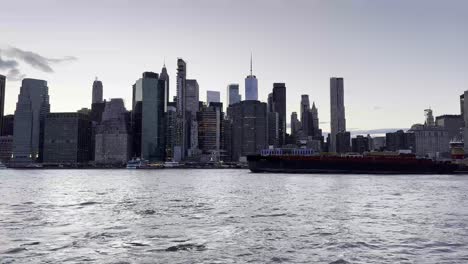 Una-Vista-Panorámica-Del-Horizonte-De-Manhattan-Al-Atardecer,-Con-El-Río-Este-En-Primer-Plano-Y-Una-Gran-Barcaza-Atravesando-El-Agua,-Reflejando-La-Grandeza-Arquitectónica-De-La-Ciudad