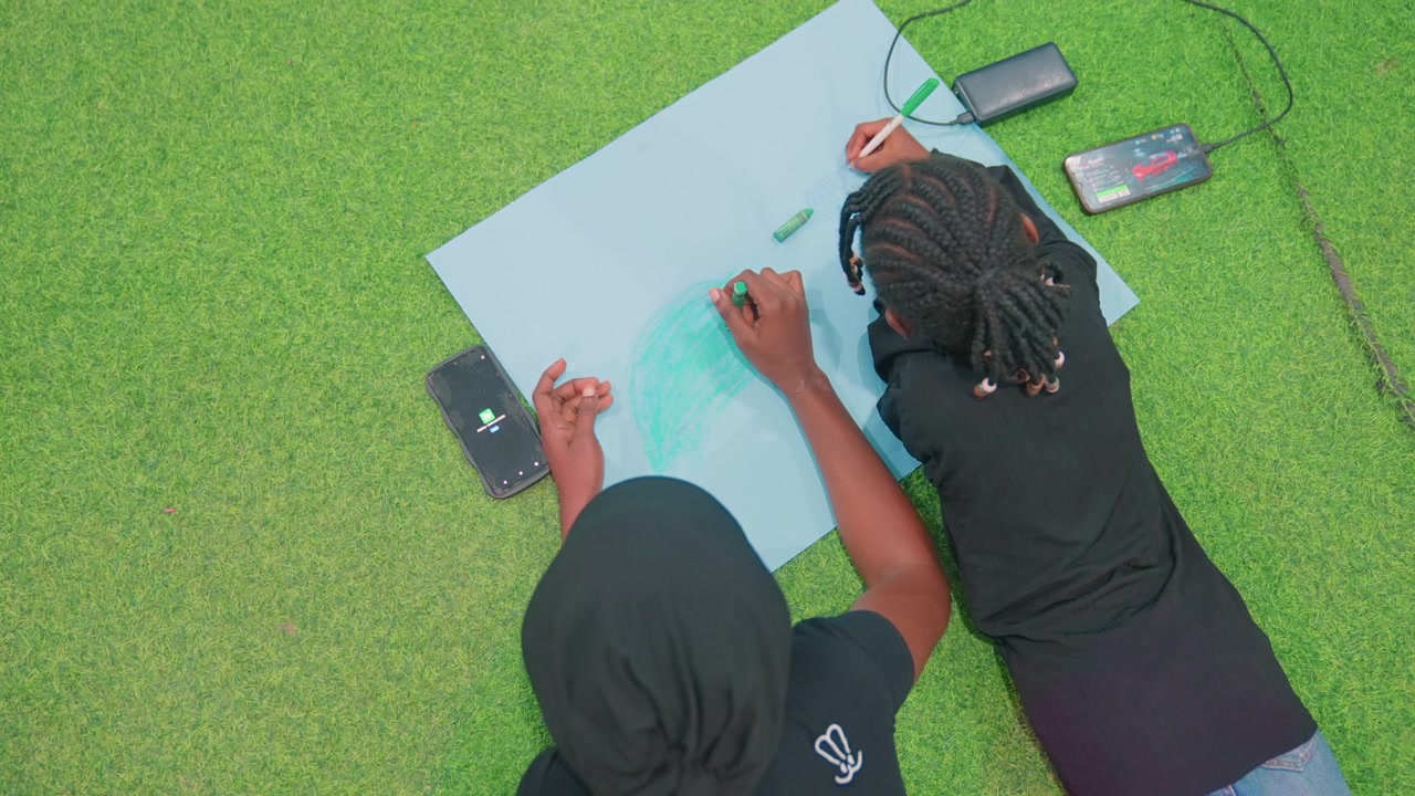Black mother and child drawing on turf, overhead view of collaborative sketching, markers and paper, smartphone reference visible, braided hair and matching black shirts, concentrated posture, mentor