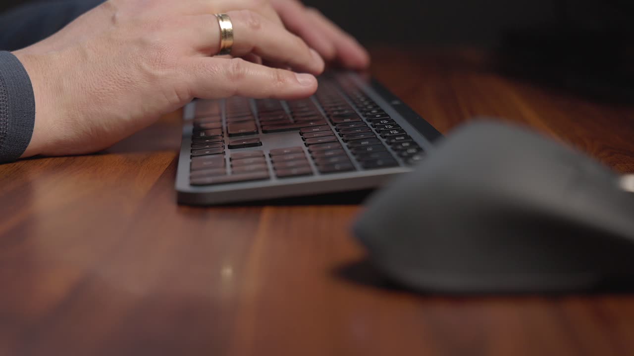 Person&rsquo;s hands wearing a grey sweater and has a ring on the left handtyping on a black keyboard placed on a polished wooden desk