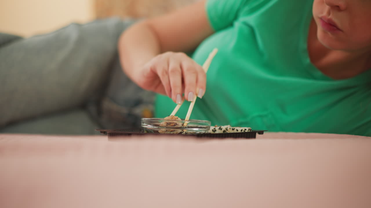 Woman eating sushi in green top lying comfortably on soft bed using chopsticks to dip sushi roll into soy sauce with soft blur background