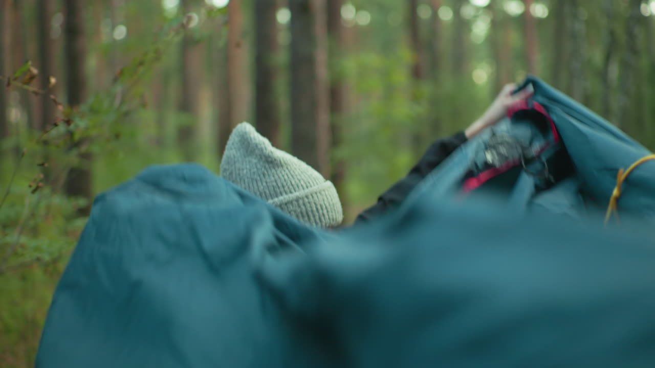 Close up of young woman in gray beanie and black jacket smiling warmly while holding tent cover in hand, engaging with companion during peaceful camping moment in green forest setting