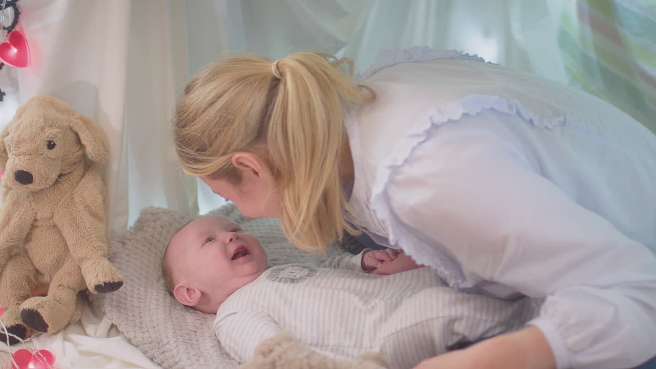Loving mother playing game with  baby son and soft toy lying on rug in homemade camp in child's bedroom at home - shot in slow motion