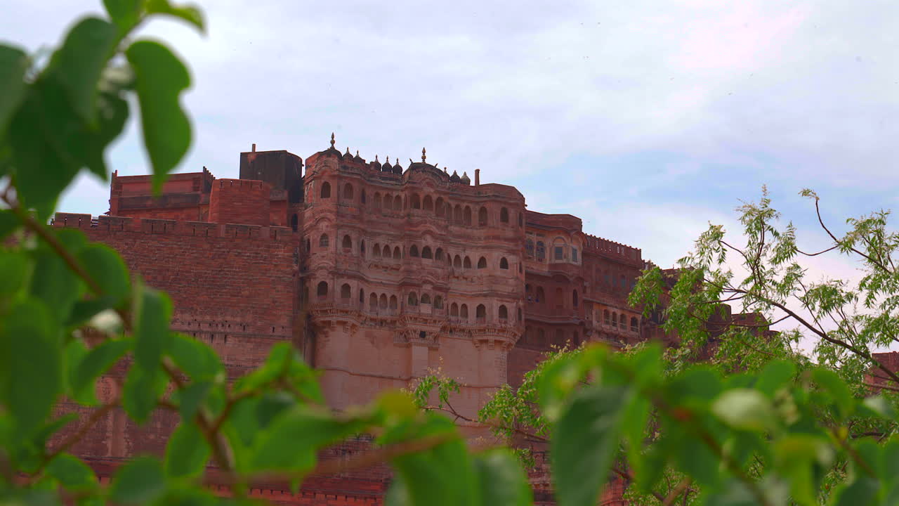 Un'inquadratura statica dell'ornata struttura superiore del palazzo del Forte Mehrangarh, incorniciata da foglie verdi in primo piano sotto un cielo coperto a Jodhpur, Rajasthan, India.
