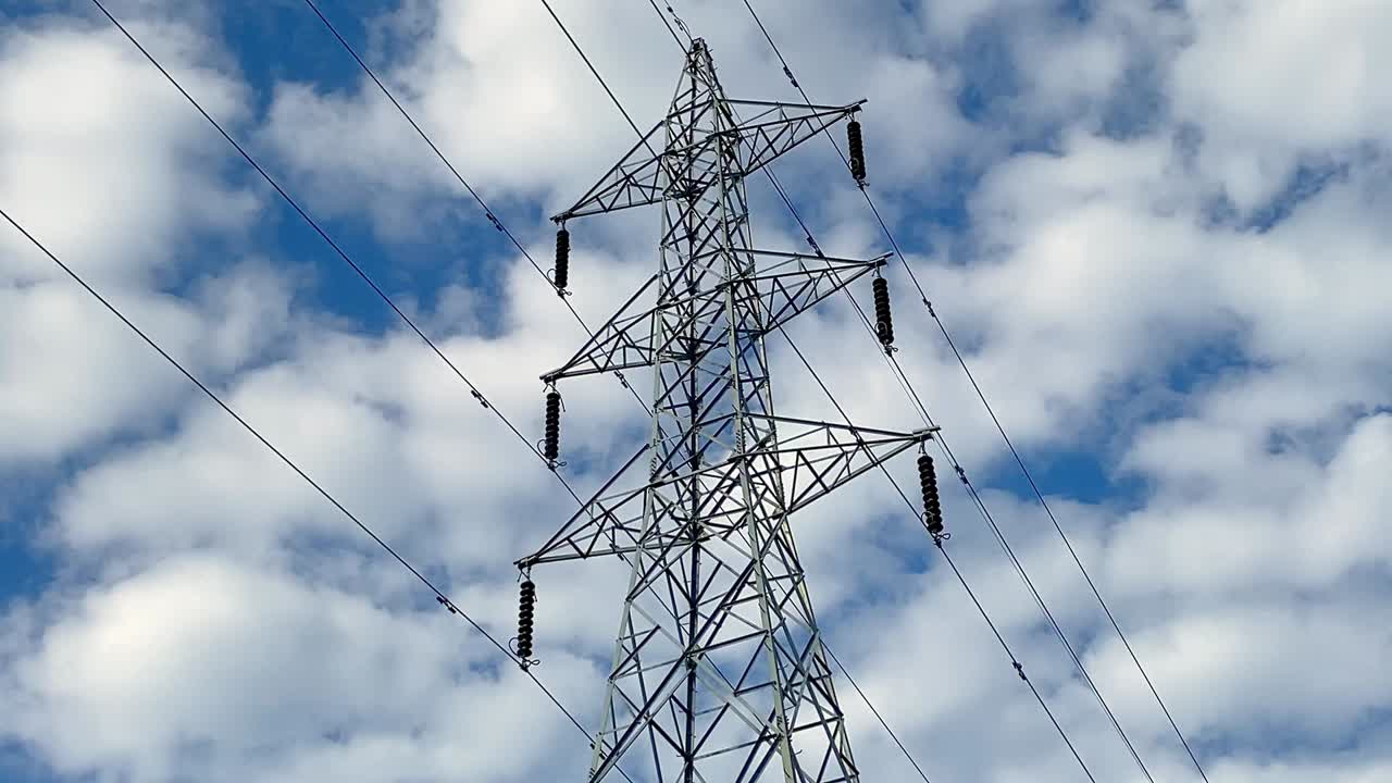High Voltage Power Lines Of A Transmission Tower Against The Blue Cloudscape Sky. Panning Up Shot