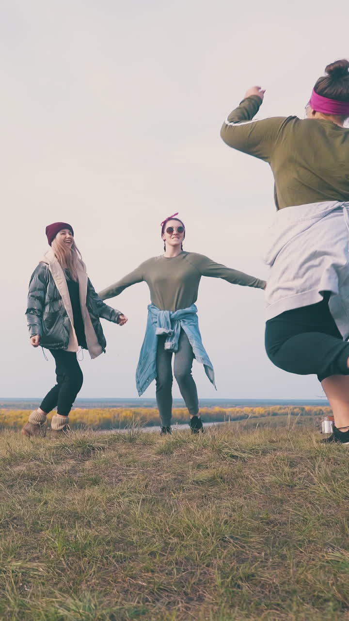 positive girls hikers dance hugging at campsite on green meadow against clear sky at sunset time low angle shot