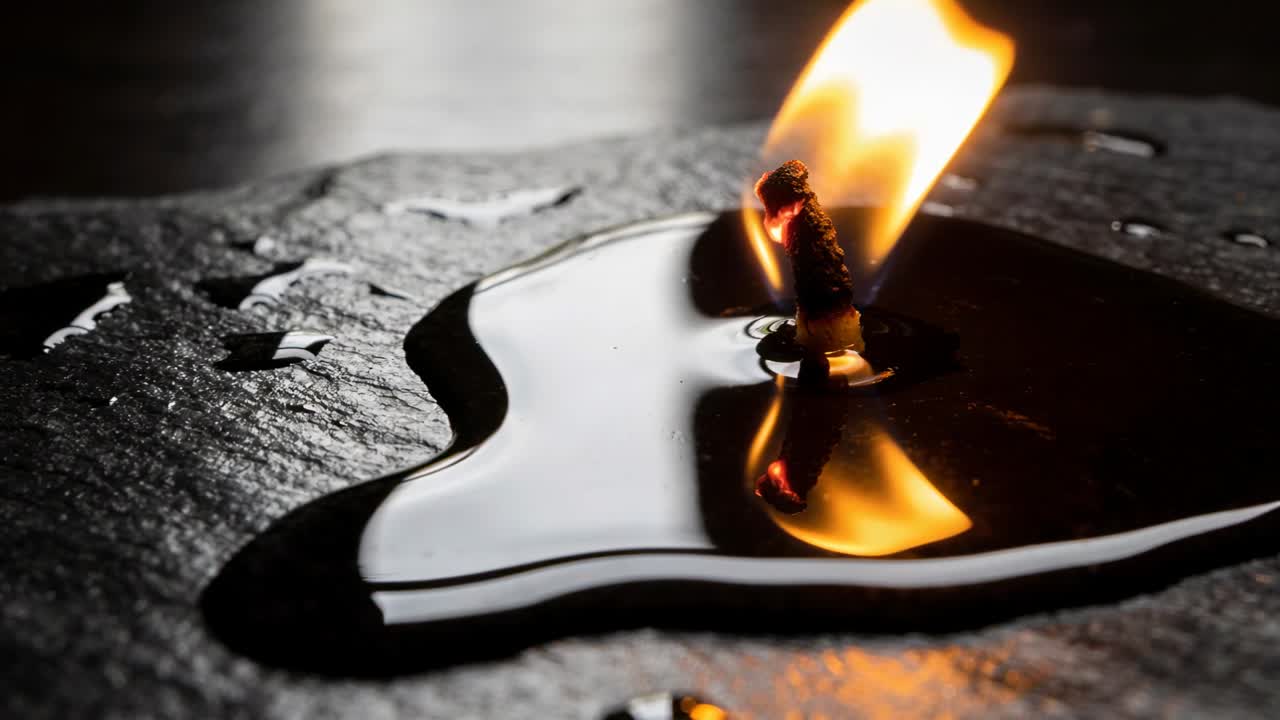 Ein angekohlter Docht entzündet sich, die Flamme verlängert sich, zieht Brennstoff aus einem flachen Pool auf Schiefer und spiegelt die Flamme.