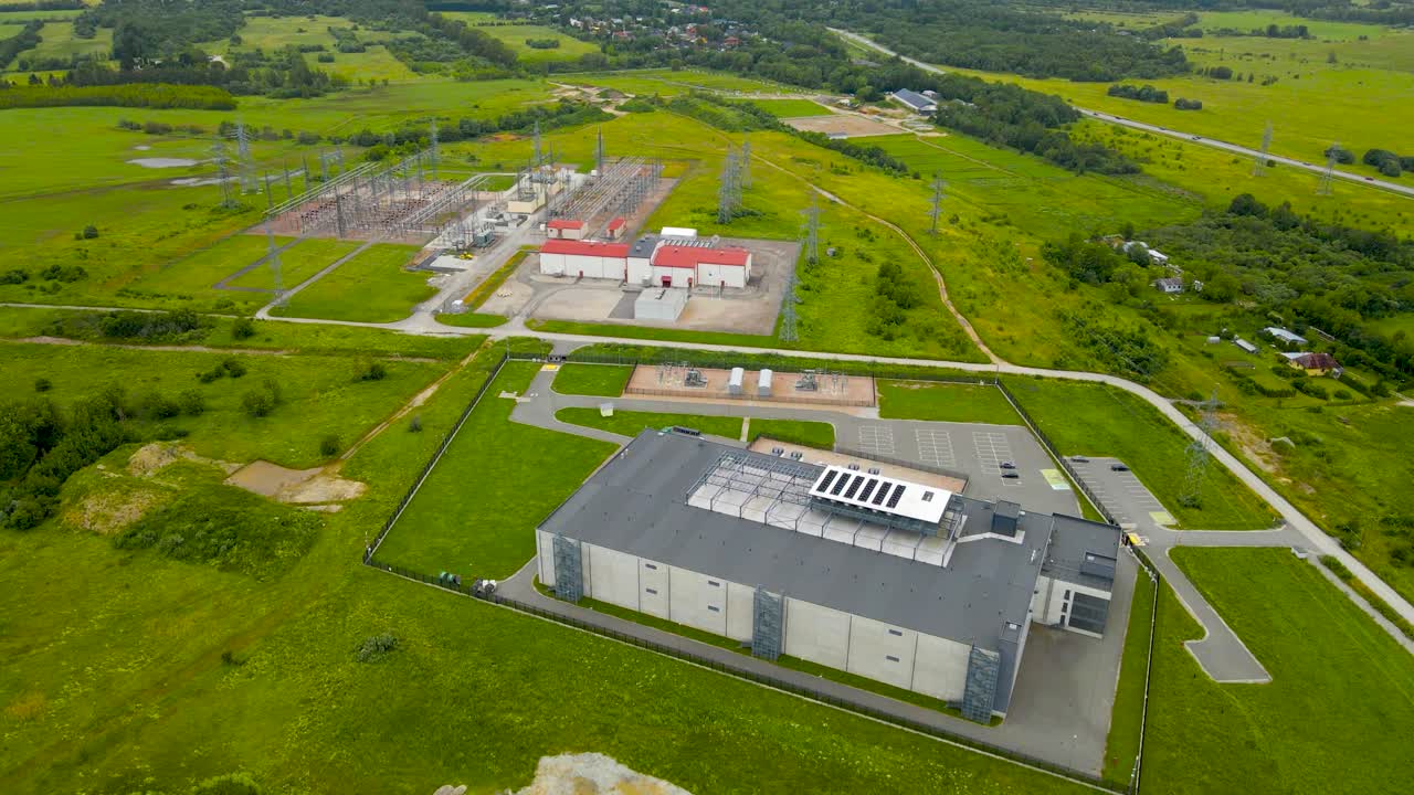 Aerial drone revealing shot of a large gray and white colored data centre with a power station that has large electrical lines next to it. The high security building is fenced and in green countryside