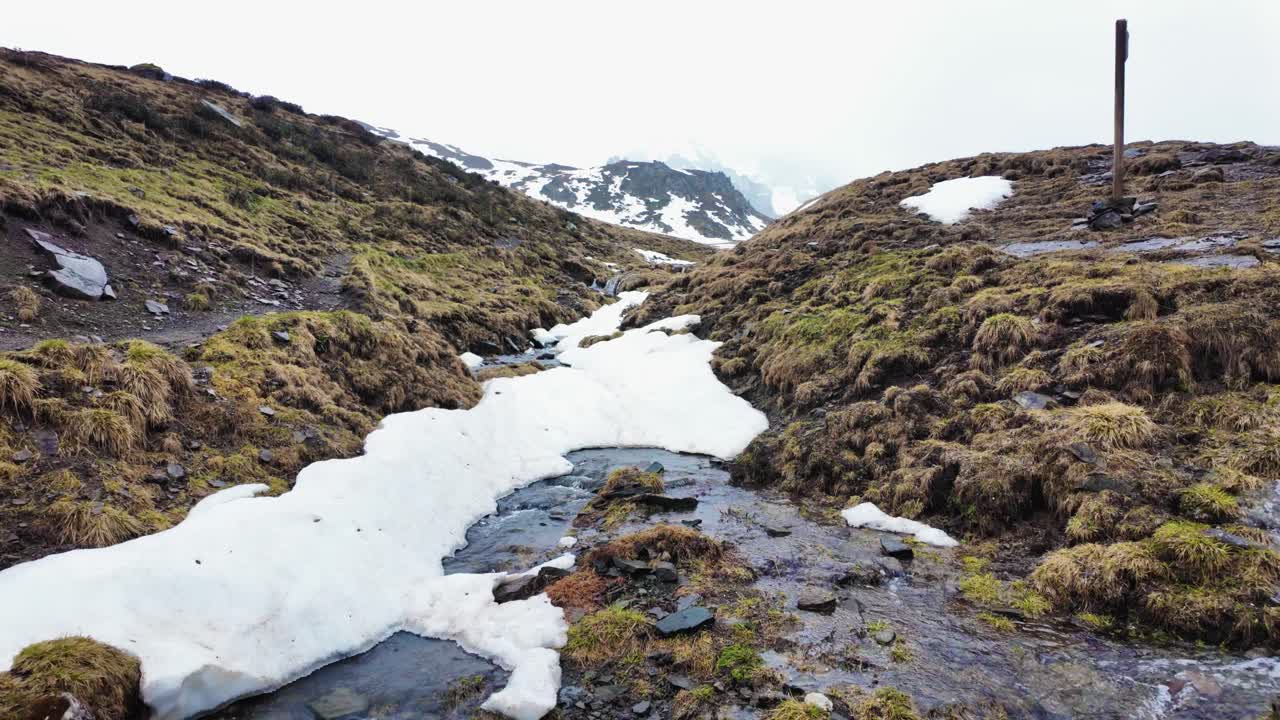Slow-motion video of Passo Giau, highlighting a melting snow stream and alpine meadow in the Italian Dolomites under an overcast sky