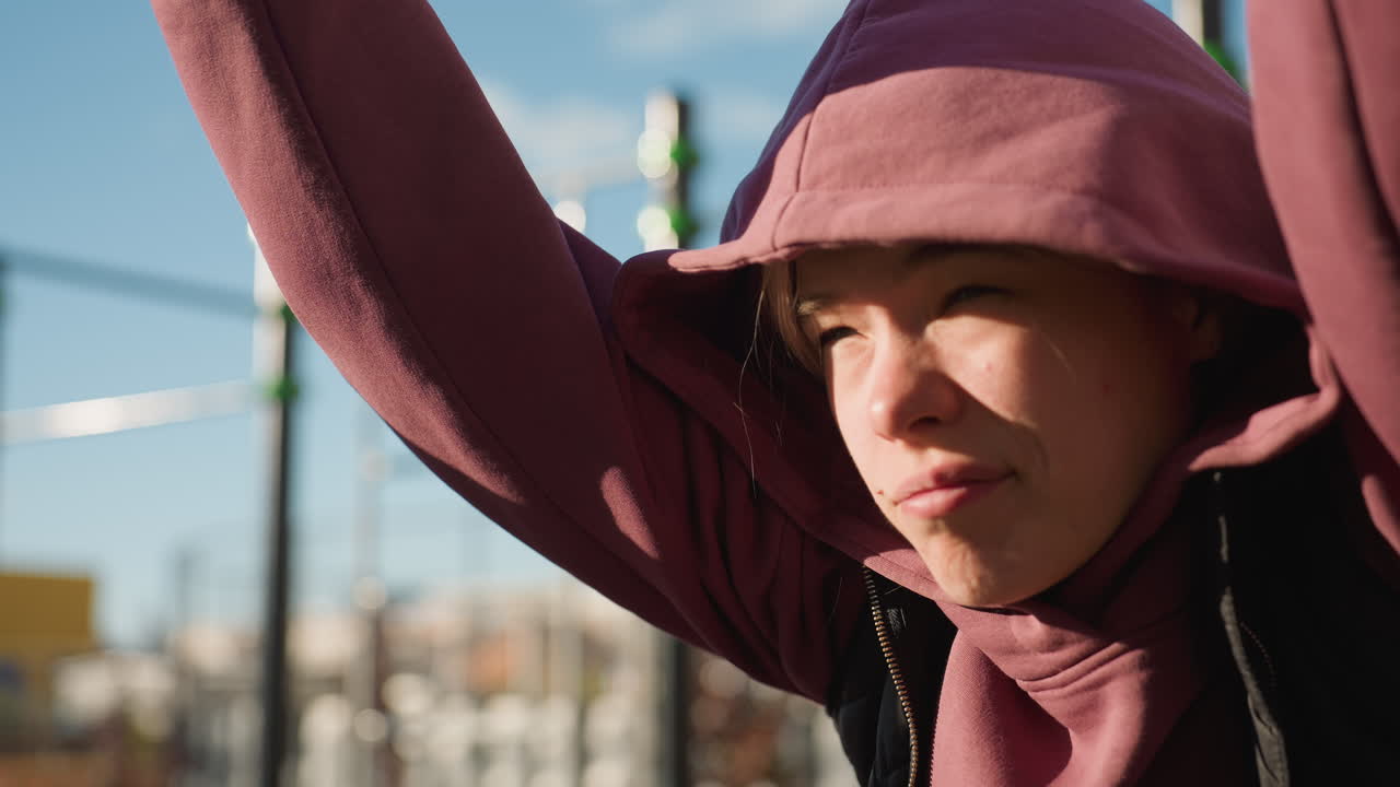 Young woman gripping metal pullup bar under bright sun hoodie clad athlete squinting against glare showing strength and resilience doing outdoor workout at urban fitness park with highrise backdrop