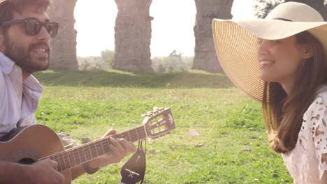 happy young lovely couple sitting lying on a blanket in the grass in front of ancient roman aqueduct ruins in parco degli acquedotti park in rome romantic play guitar sing beautiful girl with hat bucolic slow motion