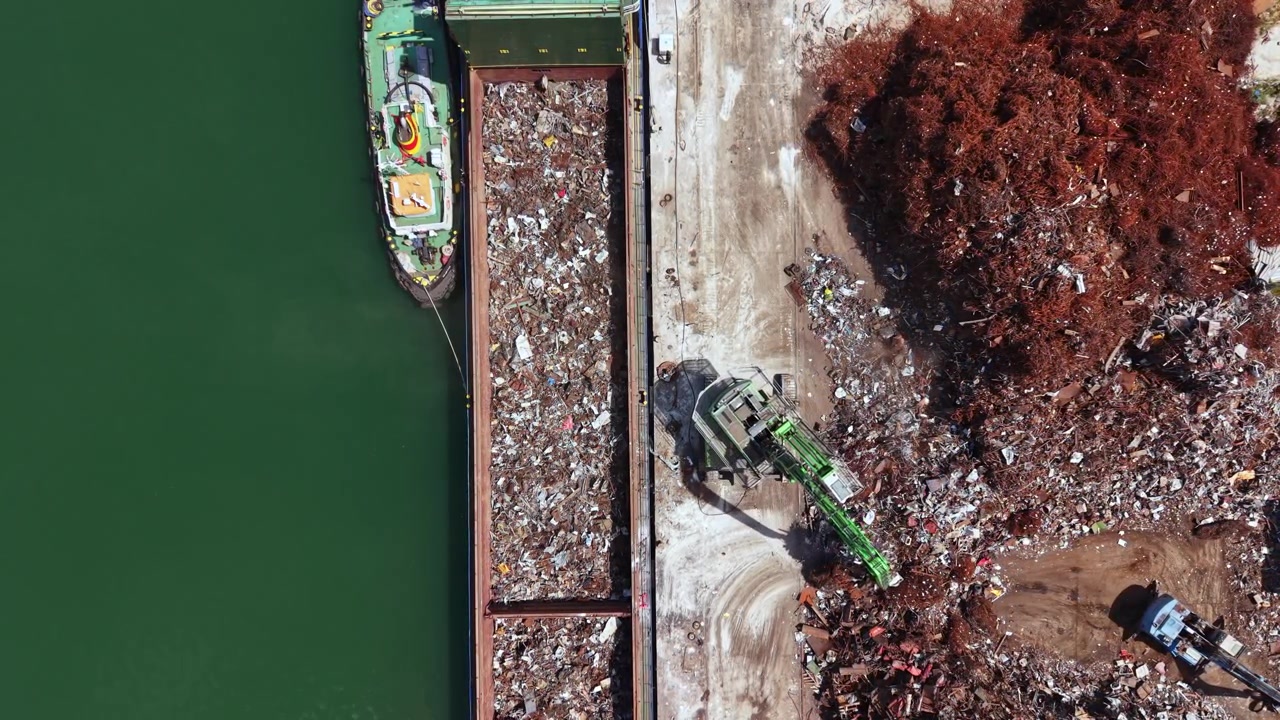 Una grande imbarcazione scarica rottami metallici a un molo mentre una gru sposta materiali vicino all'acqua durante il giorno in un'area industriale trafficata.