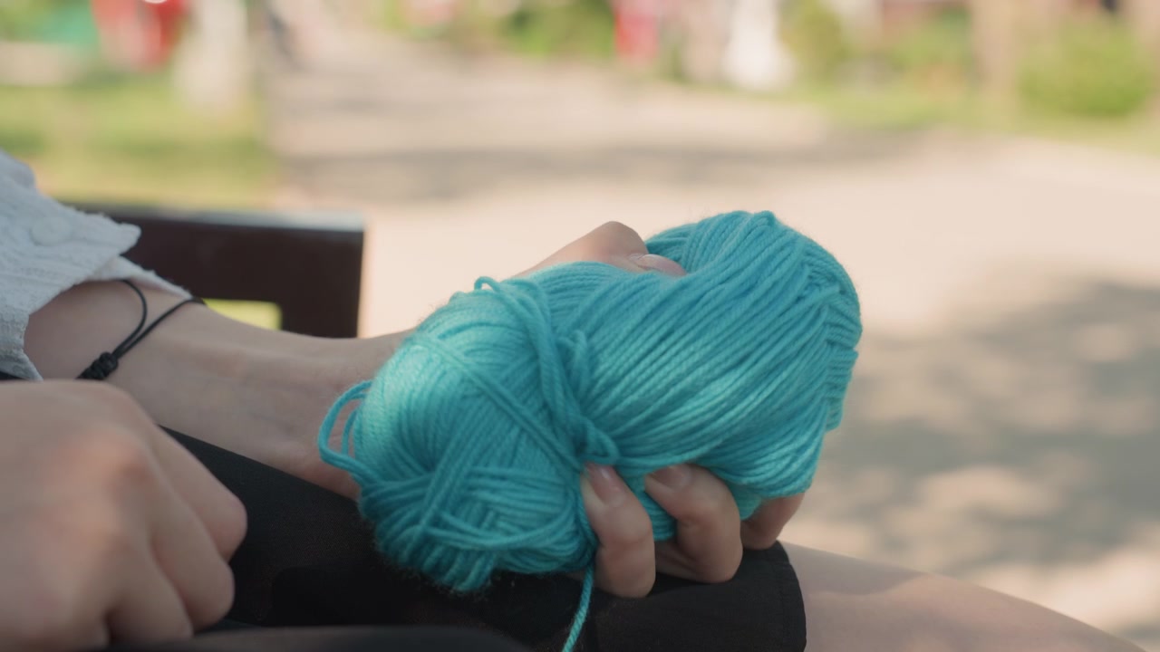 Close up of woman holding turquoise yarn in her hand while seated outdoors. focus remains on texture and color of ball of wool with soft natural light illuminating scene. Background shows