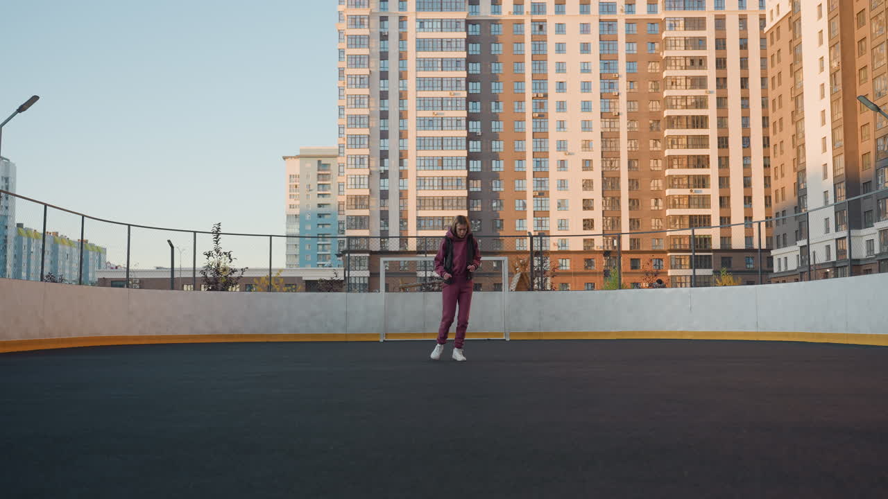 Long shot showing fitness enthusiast jogging around round outdoor court, wearing purple tracksuit and white sneakers, pumping arms under clear sky near modern tower buildings