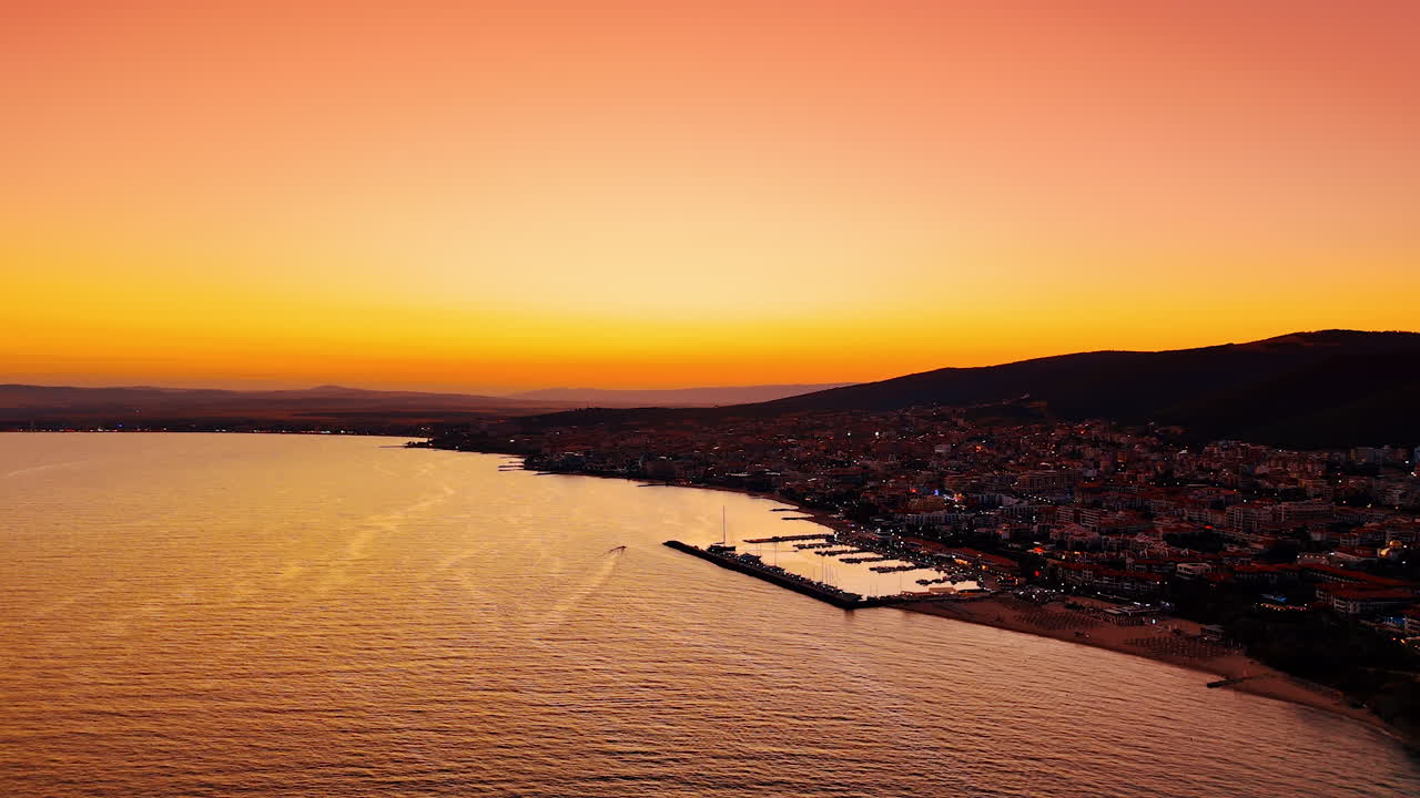 Wide aerial panorama of a coastal sunset. A soft gradient sky covers the sea and the town silhouette