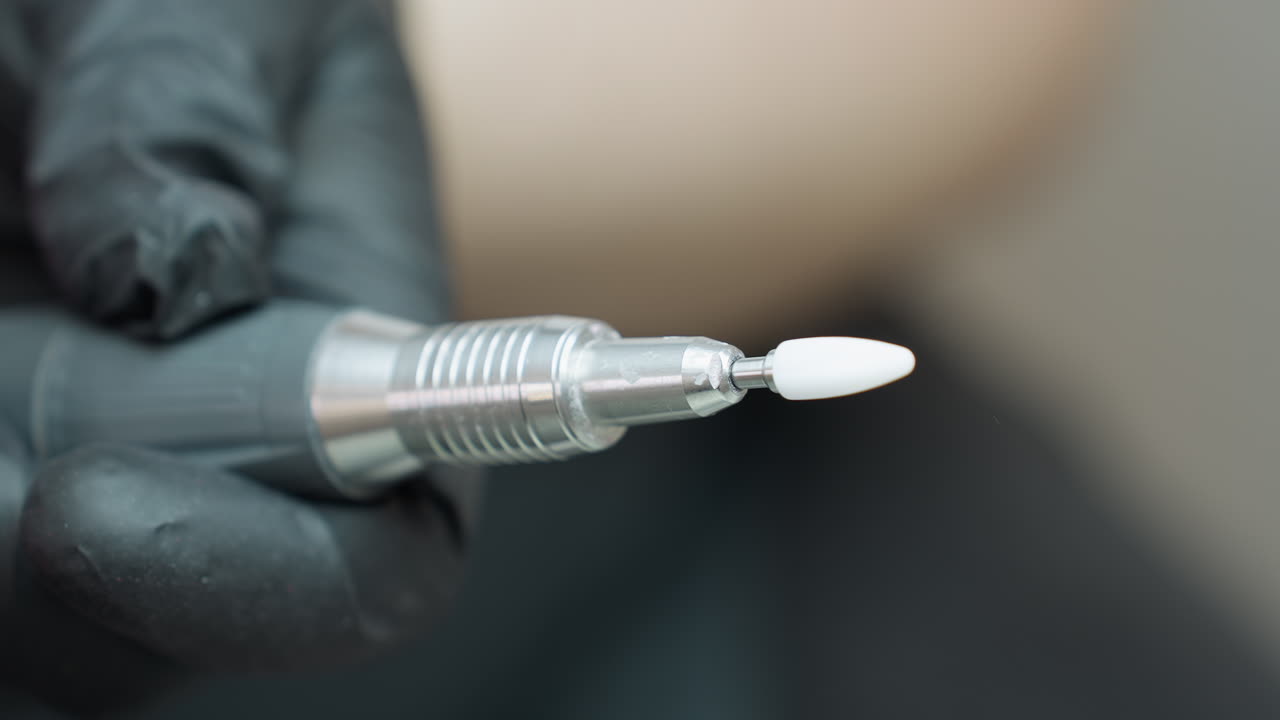 Focused view of hand wearing black glove holding silver electric nail file with white ceramic tip, emphasizing precision and hygiene in professional grooming or salon environment