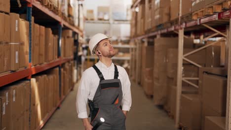 Portrait-of-a-man-in-work-uniform-with-a-flashlight-walking-down-the-warehouse-corridor-with-boxes