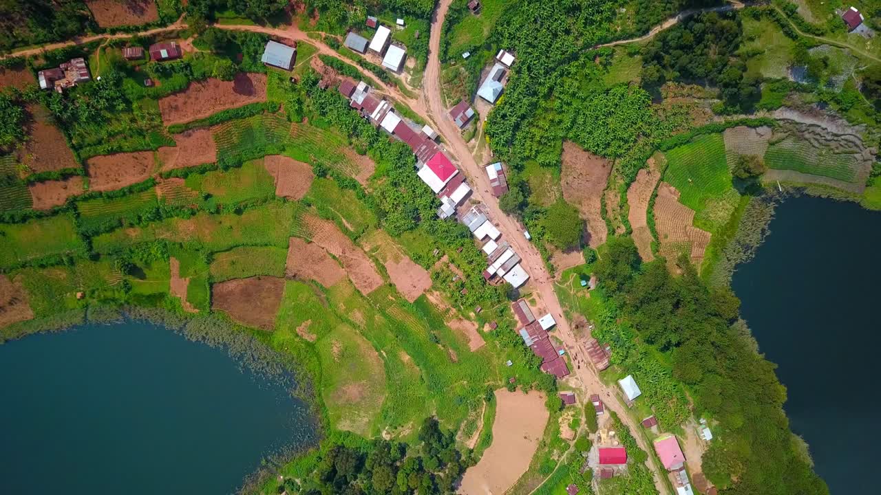Aerial shot of a rural Ugandan community with tin-roof houses atop terraced farmland between inlets of Lake Bunyonyi, showing vibrant agricultural plots, dirt roads, and traditional settlement layout