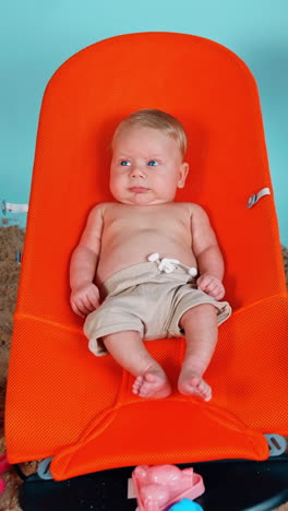 tiny infant blond boy wearing shorts lies in the orange baby chair. little kid on a beach vacation. vertical video