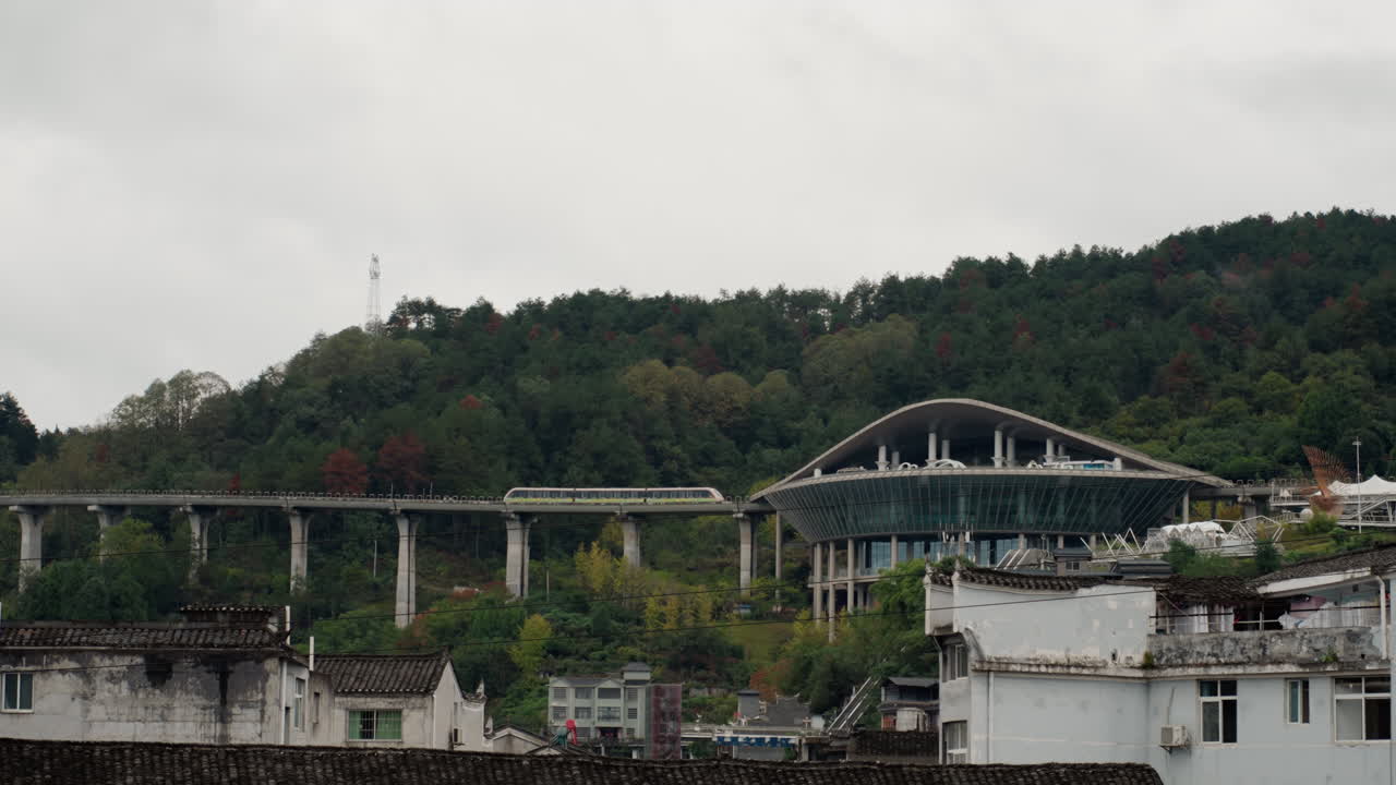 Ponte collinare sotto cielo coperto con tetti urbani, archi di viadotto in cemento che attraversano un pendio boscoso sopra vecchi edifici industriali, colori tenui, cielo nuvoloso, antenne sparse sui tetti, superfici bagnate.