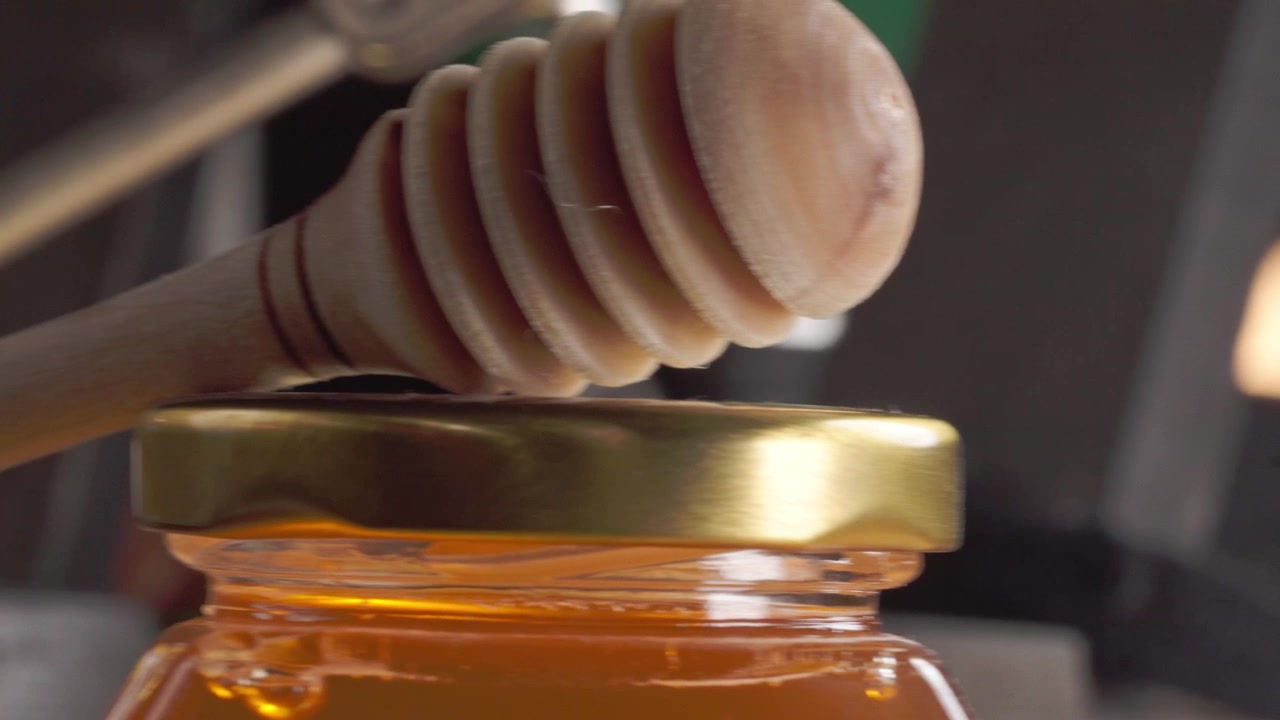 Un primo piano macro mostra un mestolo di legno per il miele posizionato sopra un barattolo di vetro pieno di miele dorato, evidenziando il liquido lucido, la texture del legno, il coperchio di metallo e i toni caldi