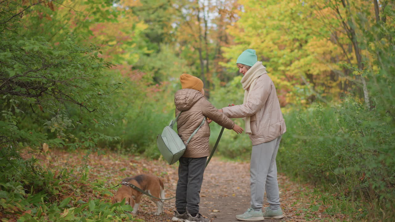 mother gently interacts with daughter while walking along forest path during autumn, both dressed warmly in jackets and hats, surrounded by lush greenery and vibrant fall foliage, dog leads ahead