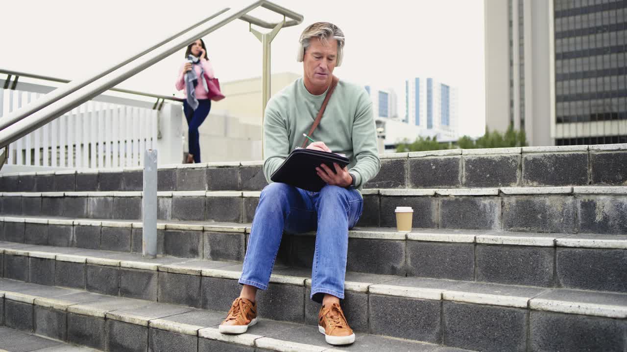Headphone-wearing mature white man tapping tablet on steps reviewing sketches with coffee cup