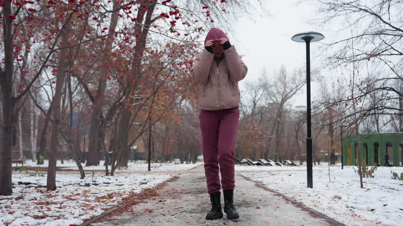 mujer con traje de invierno paseando sola, ajustando su capucha y acercando sus manos a su boca para calentarse, suelo cubierto de nieve, árboles sin hojas y hojas de otoño esparcidas