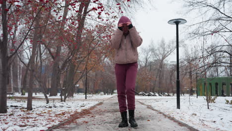 woman in winter outfit strolling alone, adjusting her hood and bringing her hands close to her mouth to warm up, snow-covered ground, leafless trees, and scattered autumn leaves