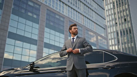 rich businessman posing car buttoning jacket. elegant man standing urban street