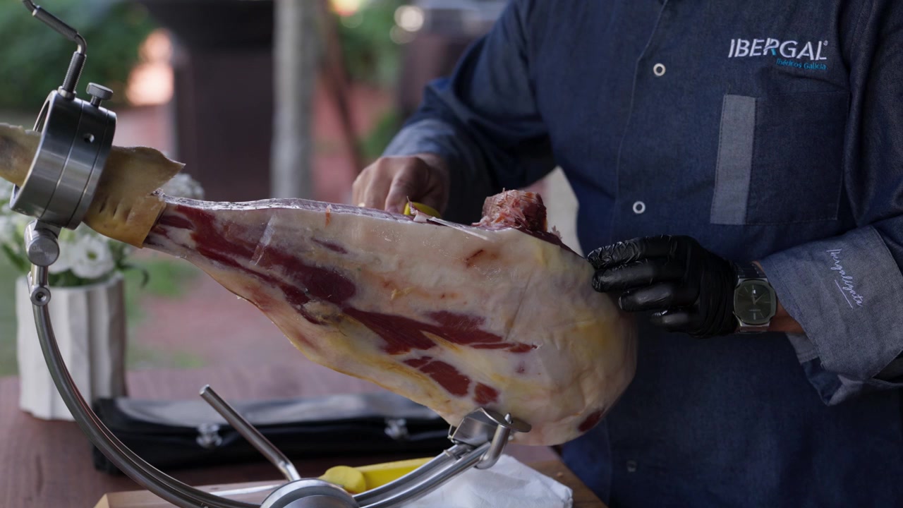 Affettatore con guanti che taglia sottili fette di prosciutto crudo da una coscia intera su un supporto all'aperto.