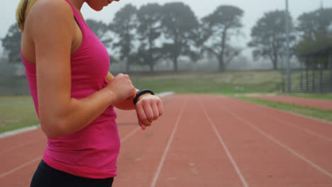 Vista-Lateral-De-Una-Atleta-Caucásica-Usando-Un-Reloj-Inteligente-En-Una-Pista-De-Atletismo-En-Un-Recinto-Deportivo-4k