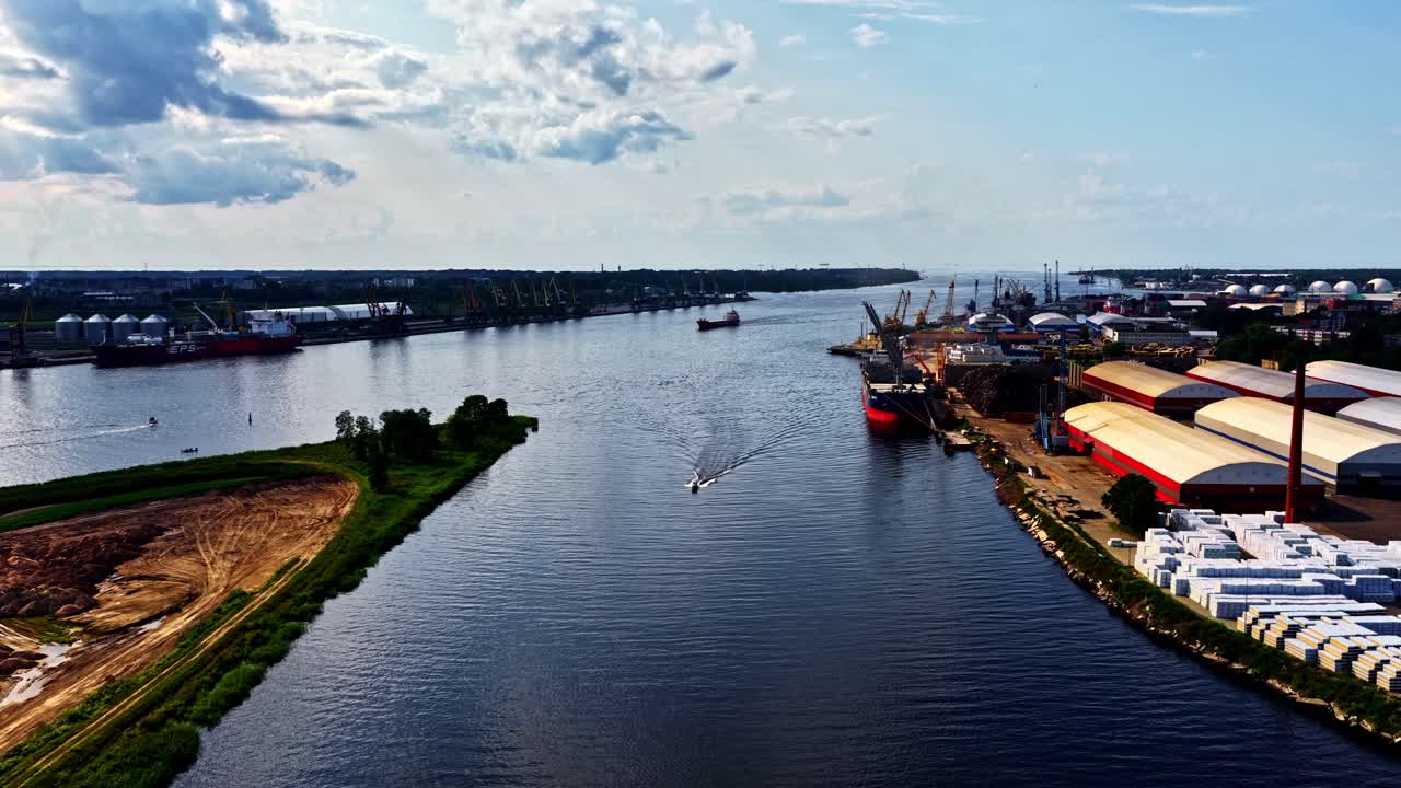 A sweeping aerial view shows the wide Daugava River flowing through the industrial Port of Riga, with cargo ships, warehouses, and cranes lining the busy riverbanks in Latvia