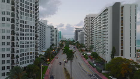 Windy-weather-in-tropical-destination.-Palm-trees-along-multilane-road-in-city-waving-in-wind.-Forwards-fly-along-modern-apartment-buildings-at-dusk.-Miami,-USA