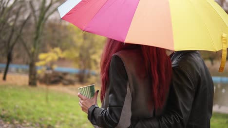 pareja feliz caminando juntos en el parque de otoño sosteniendo un paraguas colorido. hombre atractivo en chaqueta de cuero está abrazando a su novia. mujer hermosa con cabello rojo está sosteniendo una taza de papel con café