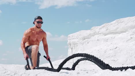 An athletic man in shorts is doing exercises with ropes on nature against the backdrop of a white mountain. Slow motion