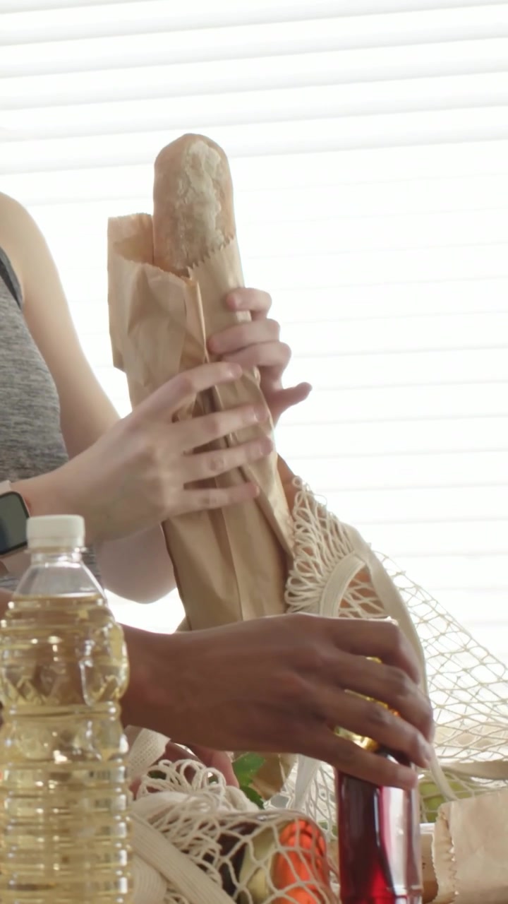 Vídeo vertical: Colocando garrafa vermelha, enquanto senhora de regata guarda baguete na sacola e desempacota na cozinha
